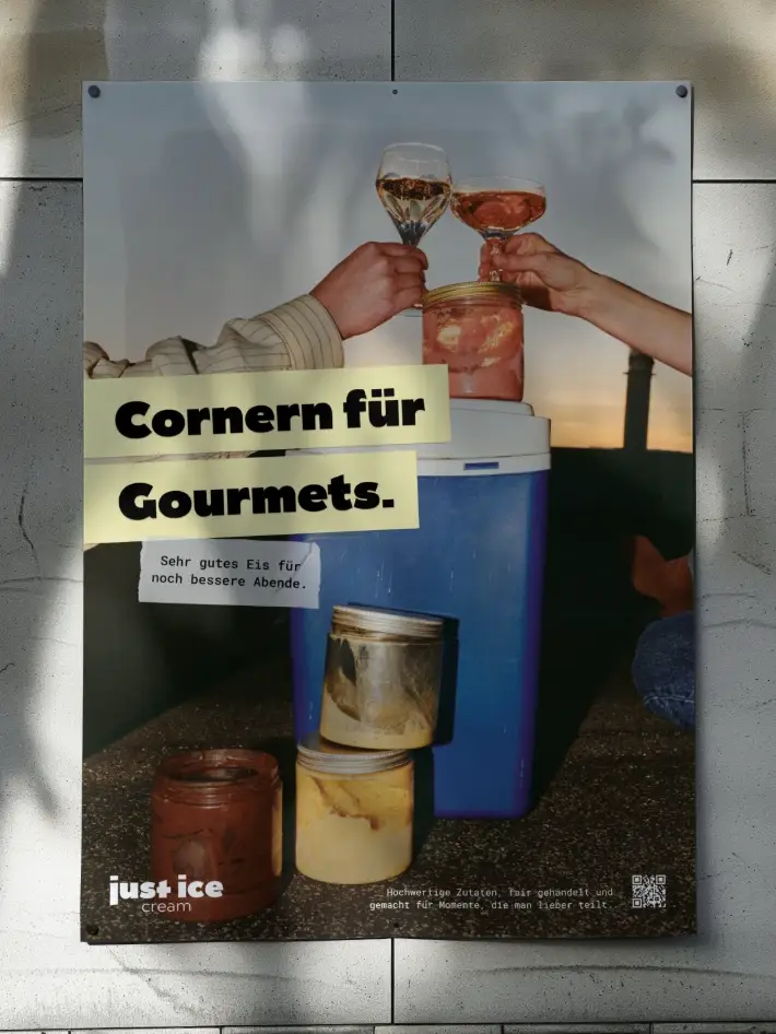 Plakat „Cornern für Gourmets.“ an einer hellen Steinwand im Licht- und Schattenspiel; das Motiv zeigt zwei anstoßende Gläser über einer Kühlbox mit gestapelten Eisgläsern.