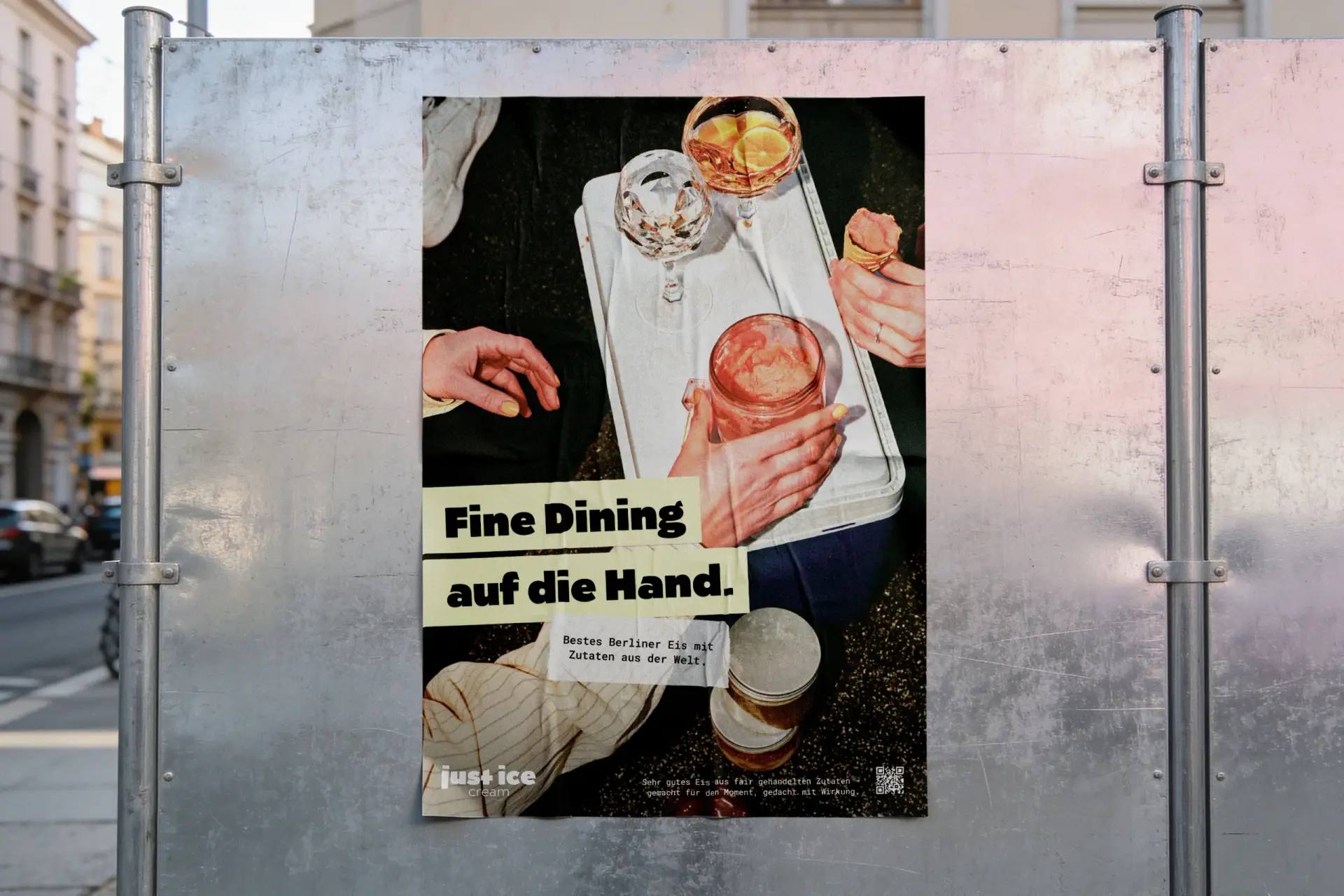 Plakat „Fine Dining auf die Hand.“ auf einer Metallwand am Straßenrand, im Hintergrund Altbauten und parkende Autos.