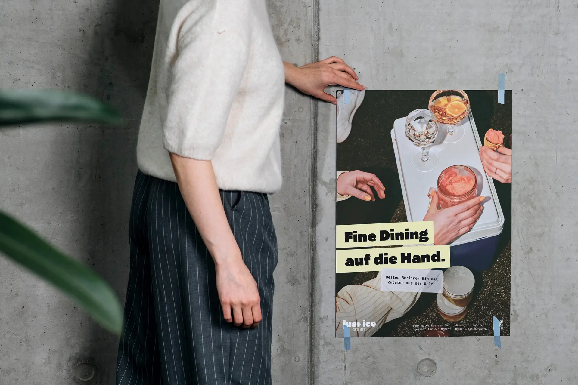 Plakat „Fine Dining auf die Hand.“ an einer Betonwand, daneben eine Person im Strickpullover; das Motiv zeigt Hände, Gläser und ein Eisglas auf einer Kühlbox.